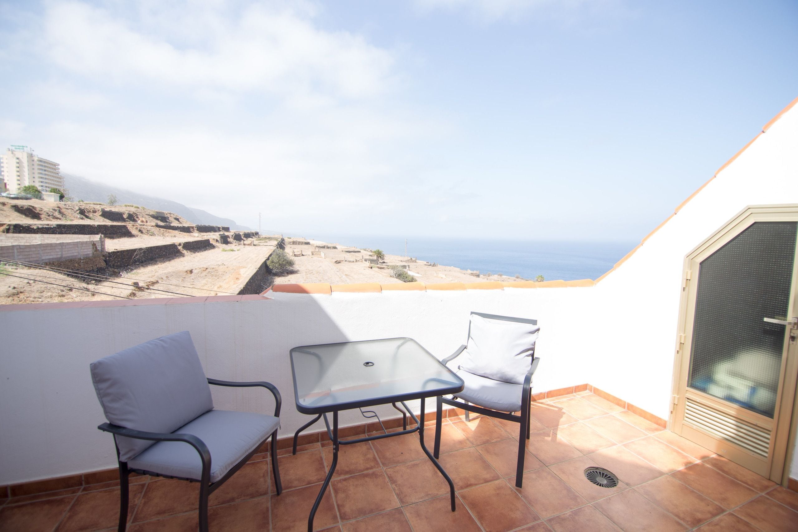 A balcony with two chairs and a table, overlooking a coastal landscape with buildings and a clear sky.