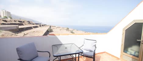 A balcony with two chairs and a table, overlooking a coastal landscape with buildings and a clear sky.