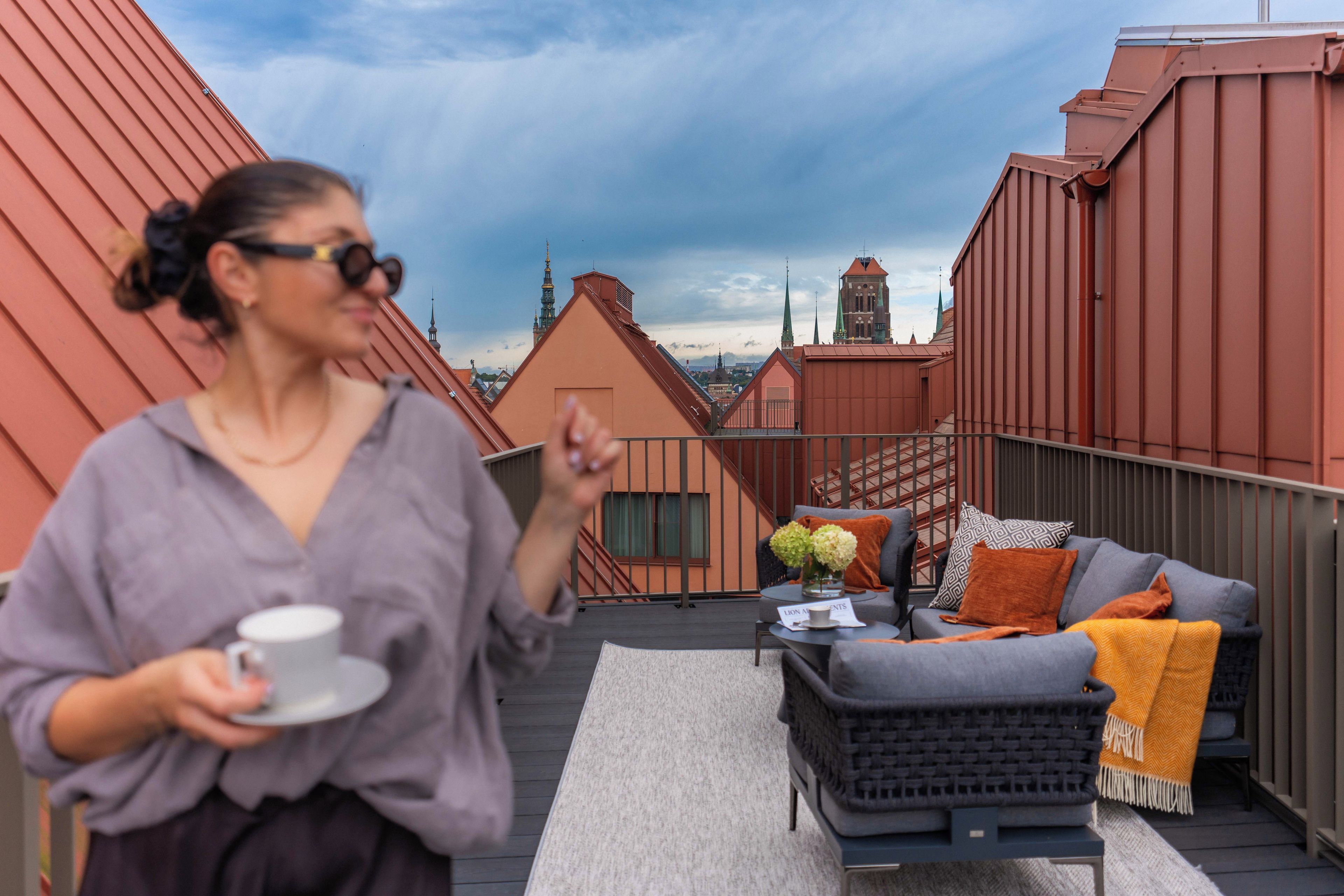 A person on a rooftop terrace with a sofa, cushions, and a table with a cup and saucer.