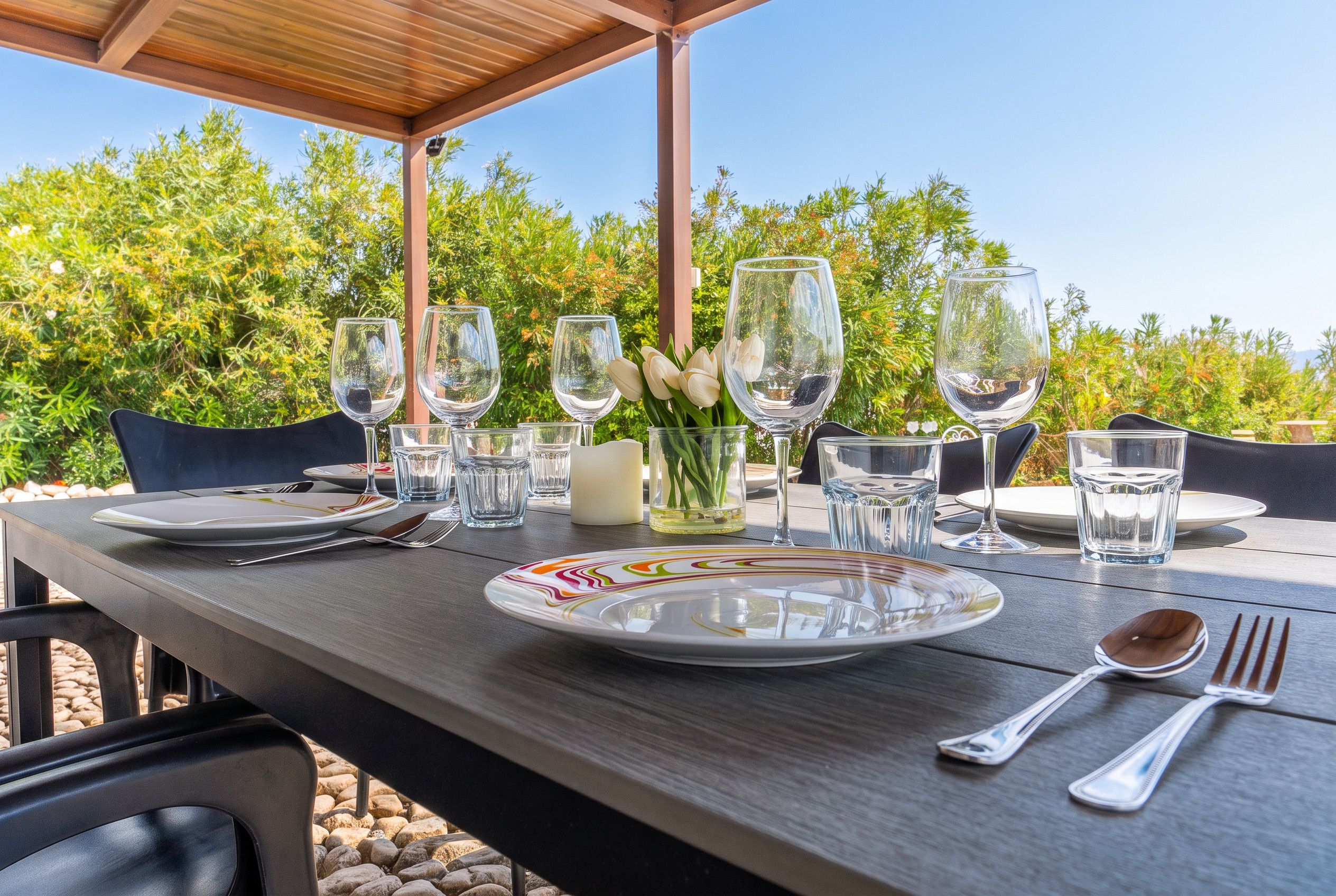 An outdoor dining table set with plates, cutlery, and wine glasses, with a view of greenery in the background.