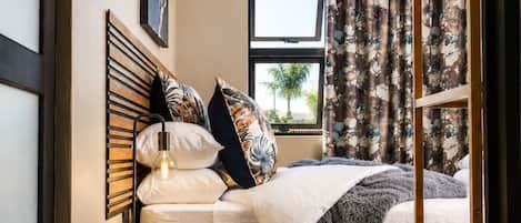 Un dormitorio con cama, mesitas de noche, una ventana con cortinas y vista a palmeras.
