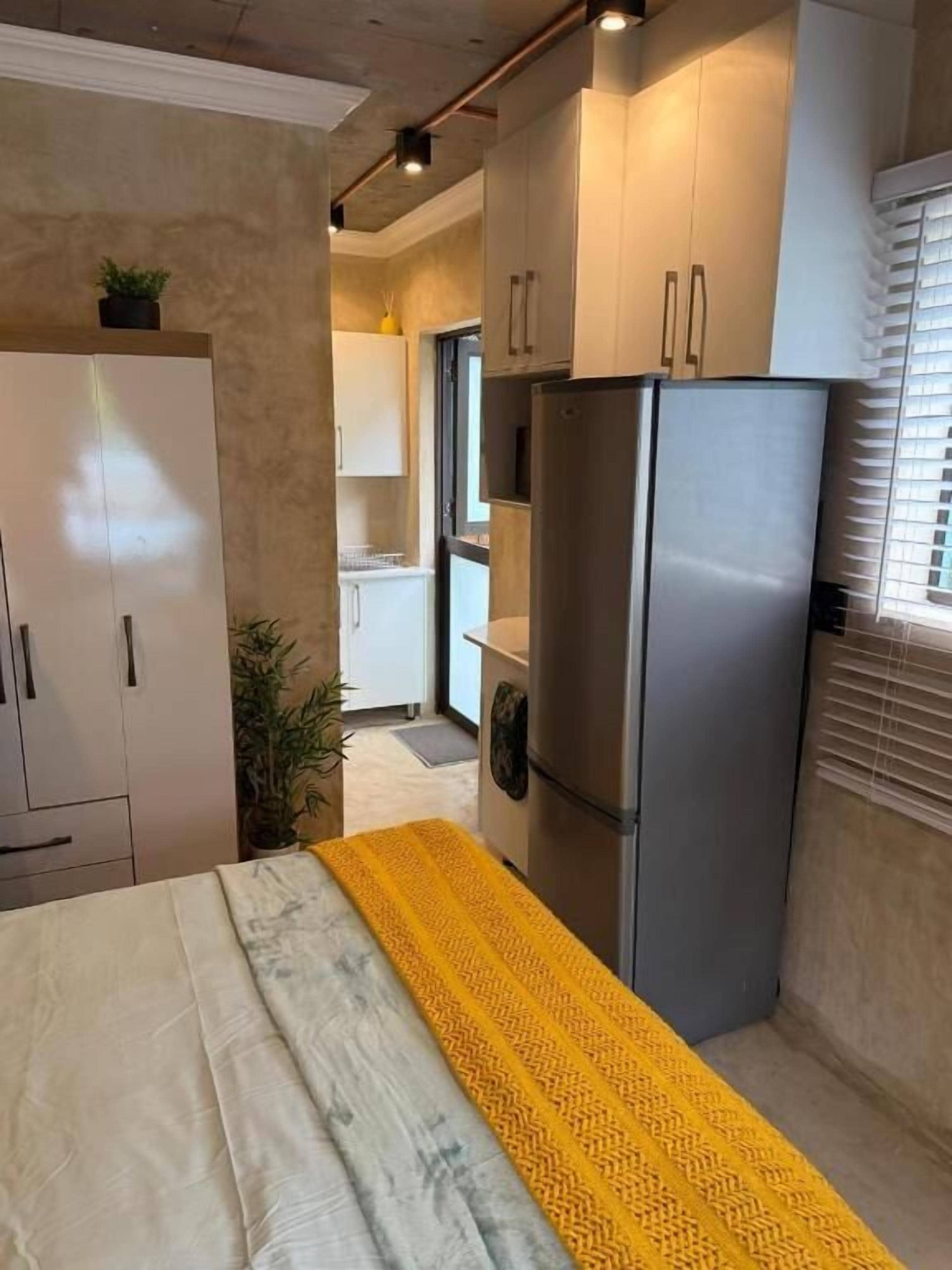 Una cocina compacta con refrigerador, armarios y una cama con colcha estampada.