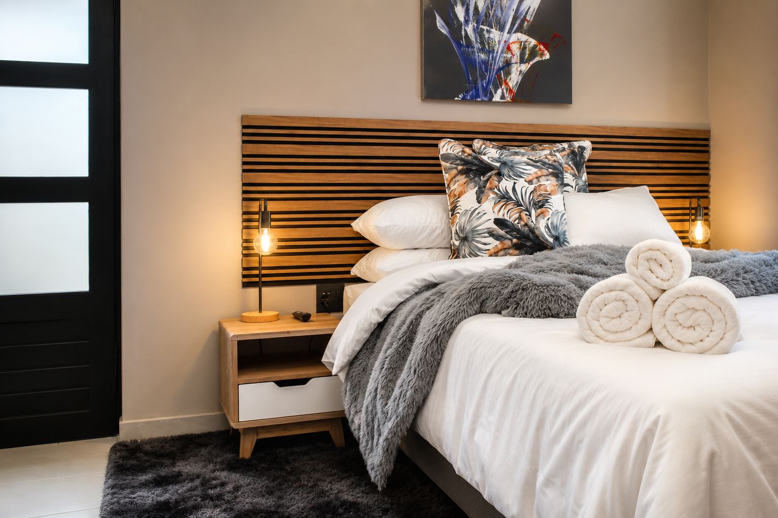 Un dormitorio con cama, mesita de noche, lámpara y una obra de arte en la pared.