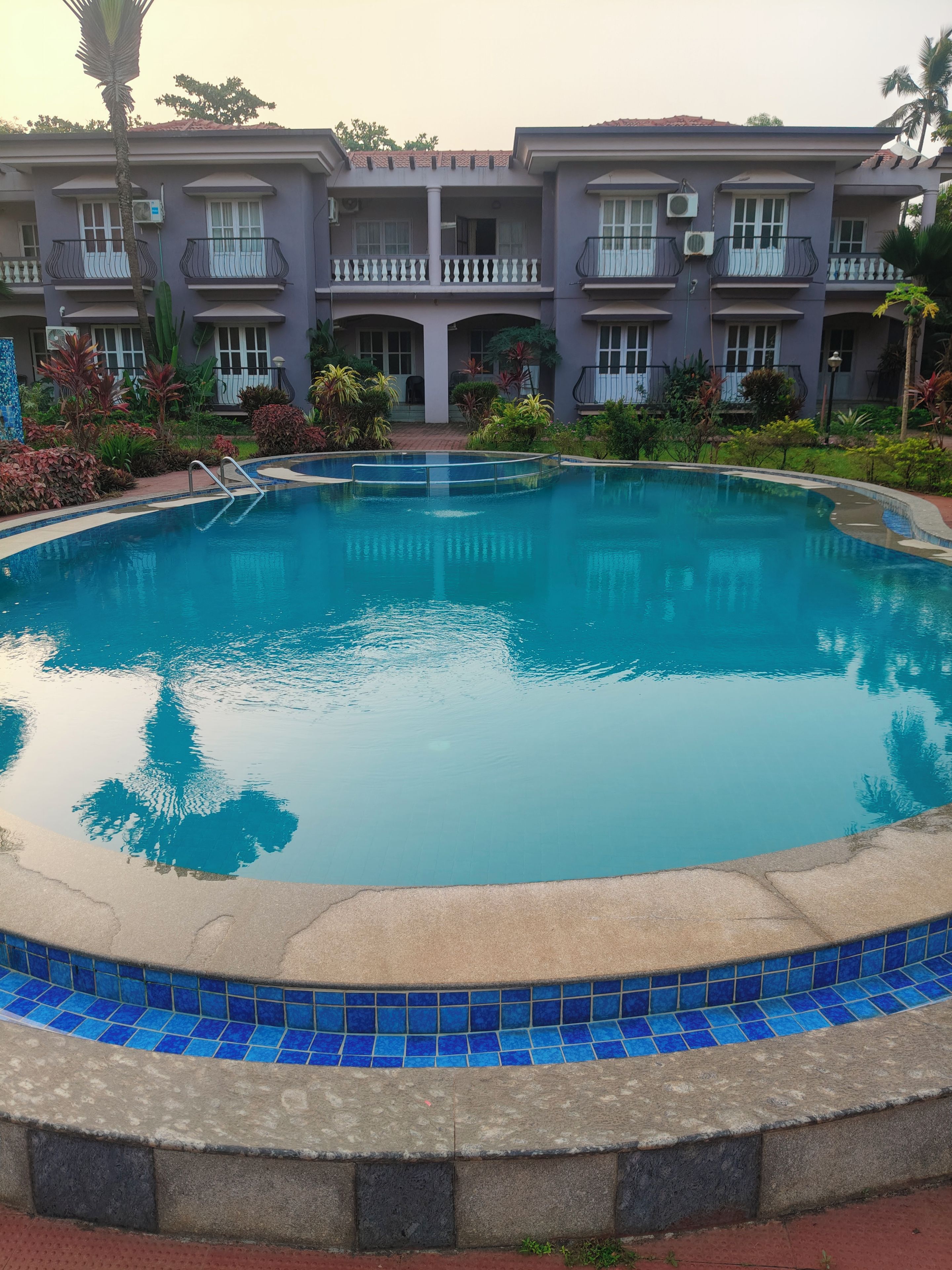 A swimming pool with a tiled edge, surrounded by a landscaped garden and a multi-story building with balconies.
