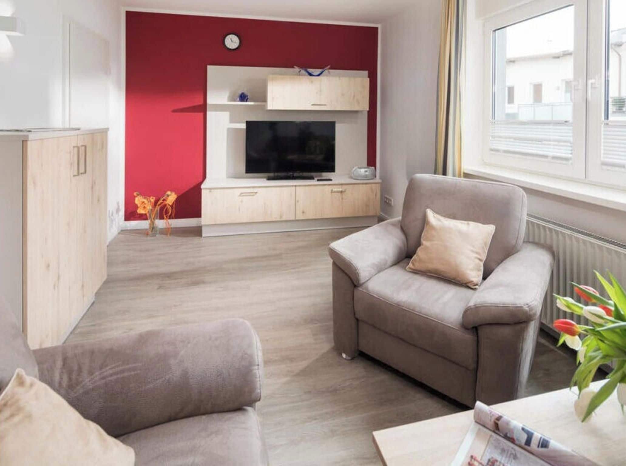 Una sala de estar con una pared de acento roja, un televisor de pantalla plana, un sofá gris y una ventana con persianas.