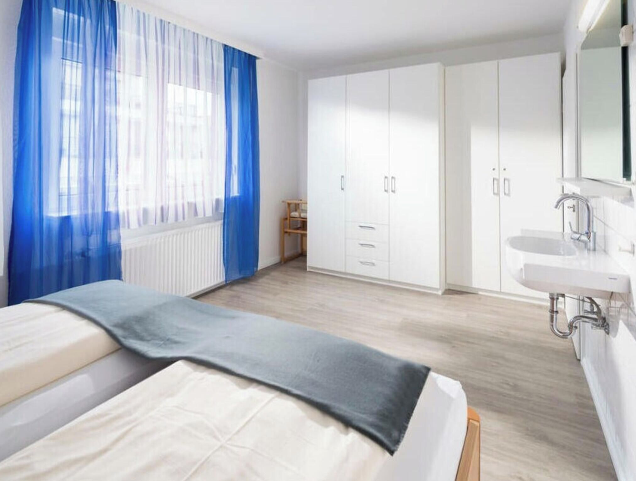 A bedroom with two beds, a wardrobe, a bathroom with a mirror, and a window with blue curtains.