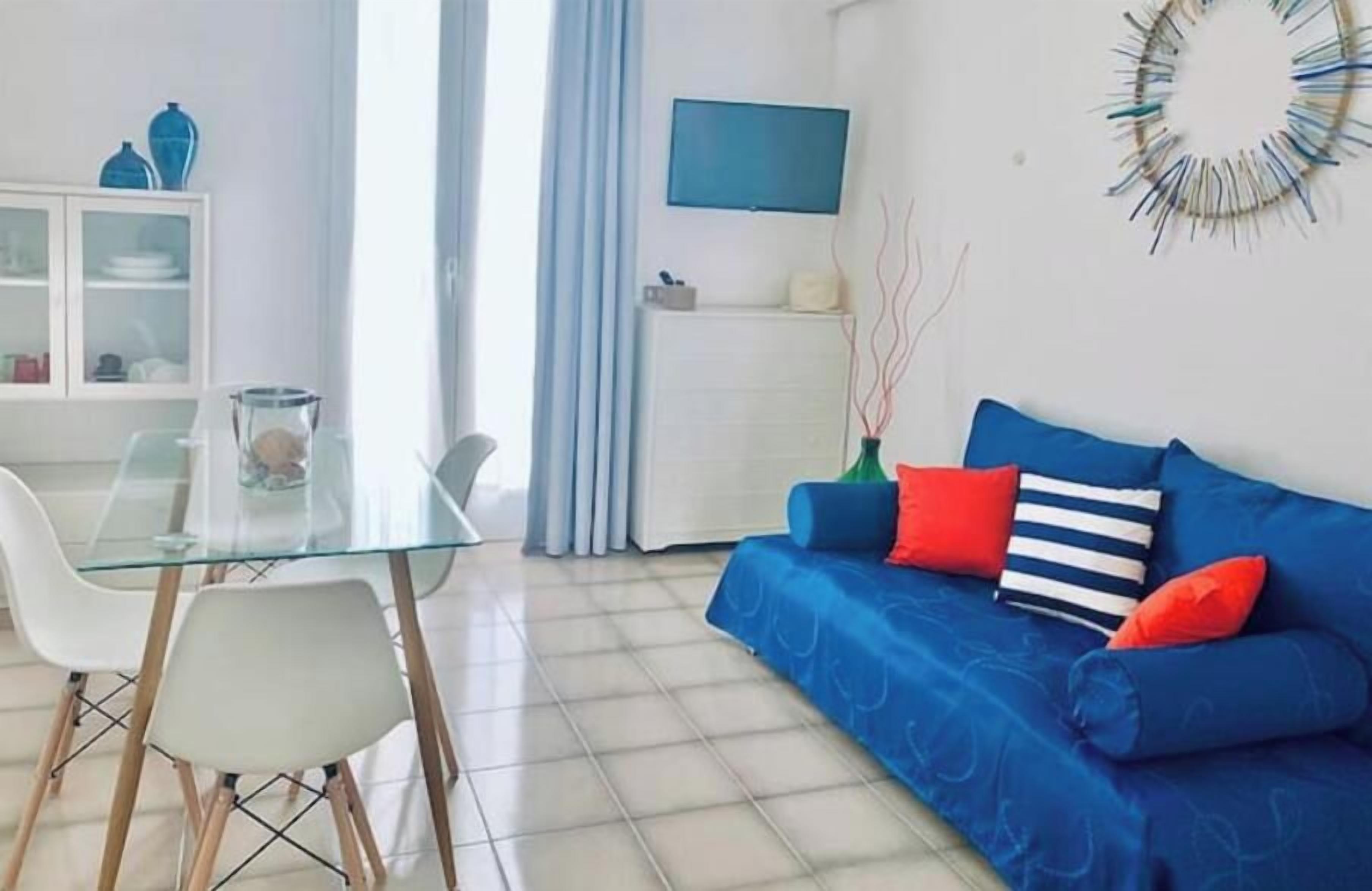 Ein modernes Wohnzimmer mit einem Glastisch, weißen Stühlen, einem blauen Sofa mit roten Kissen, einer weißen Kommode und einem Wand-Fernseher.