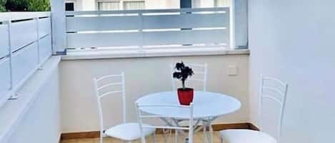 Un pequeño balcón exterior con una mesa y sillas, una planta en maceta y vistas a edificios al fondo.