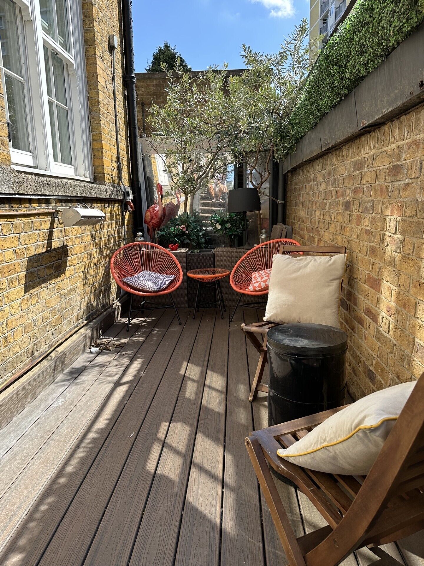 Une terrasse en bois, avec des chaises et une table en osier, entourée de murs en briques et d’un arbre.