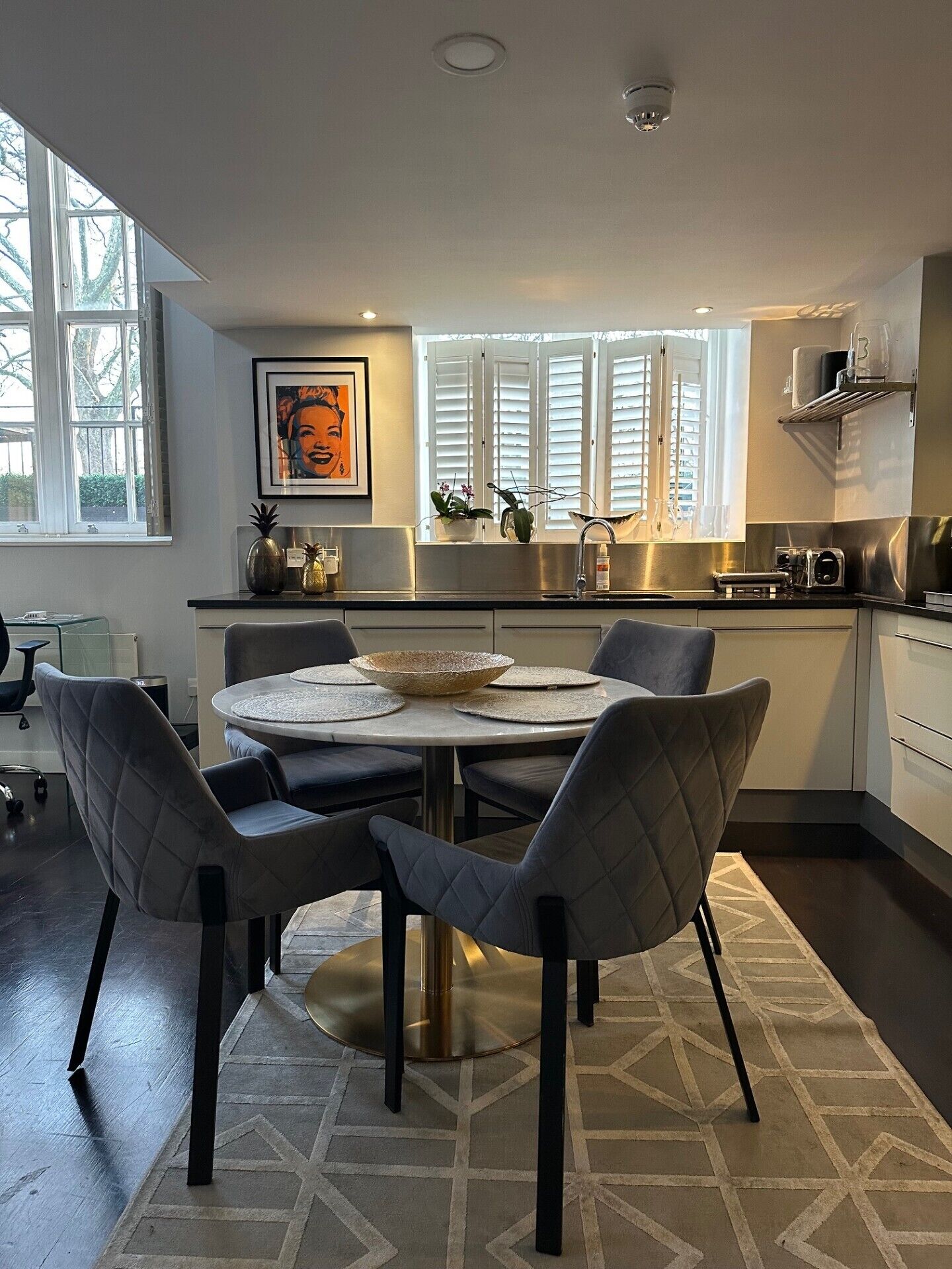 A modern kitchen with a dining area, featuring a round table with chairs, a window with blinds, and a kitchen counter with appliances.