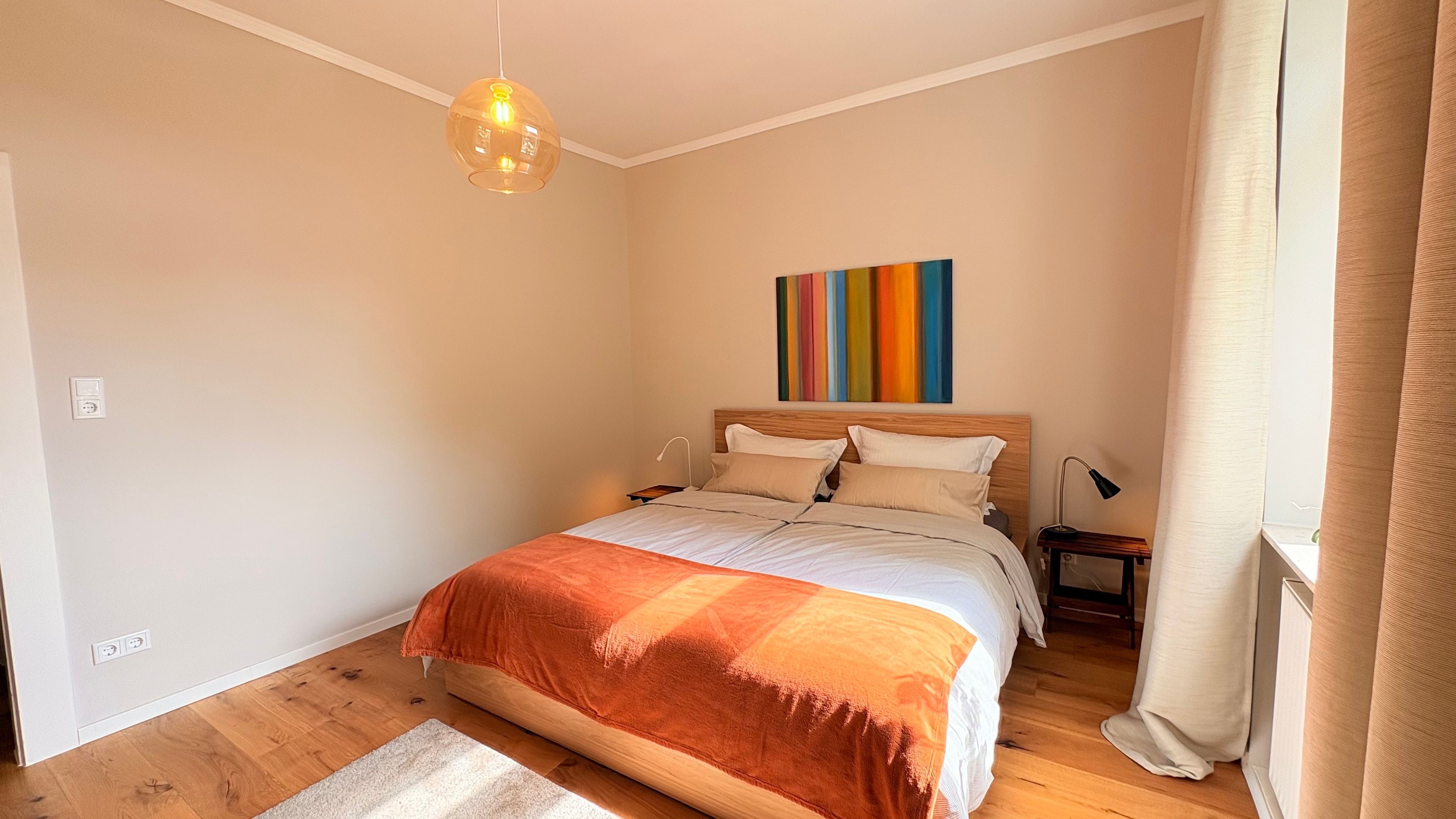 Ein Schlafzimmer mit einem Bett, einem Nachttisch, einer Lampe, einem Wandbild und einem Fenster mit Vorhängen.