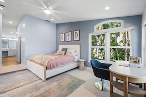 A bedroom with a bed, a desk, a chair, a window with a view of greenery, and a ceiling fan.