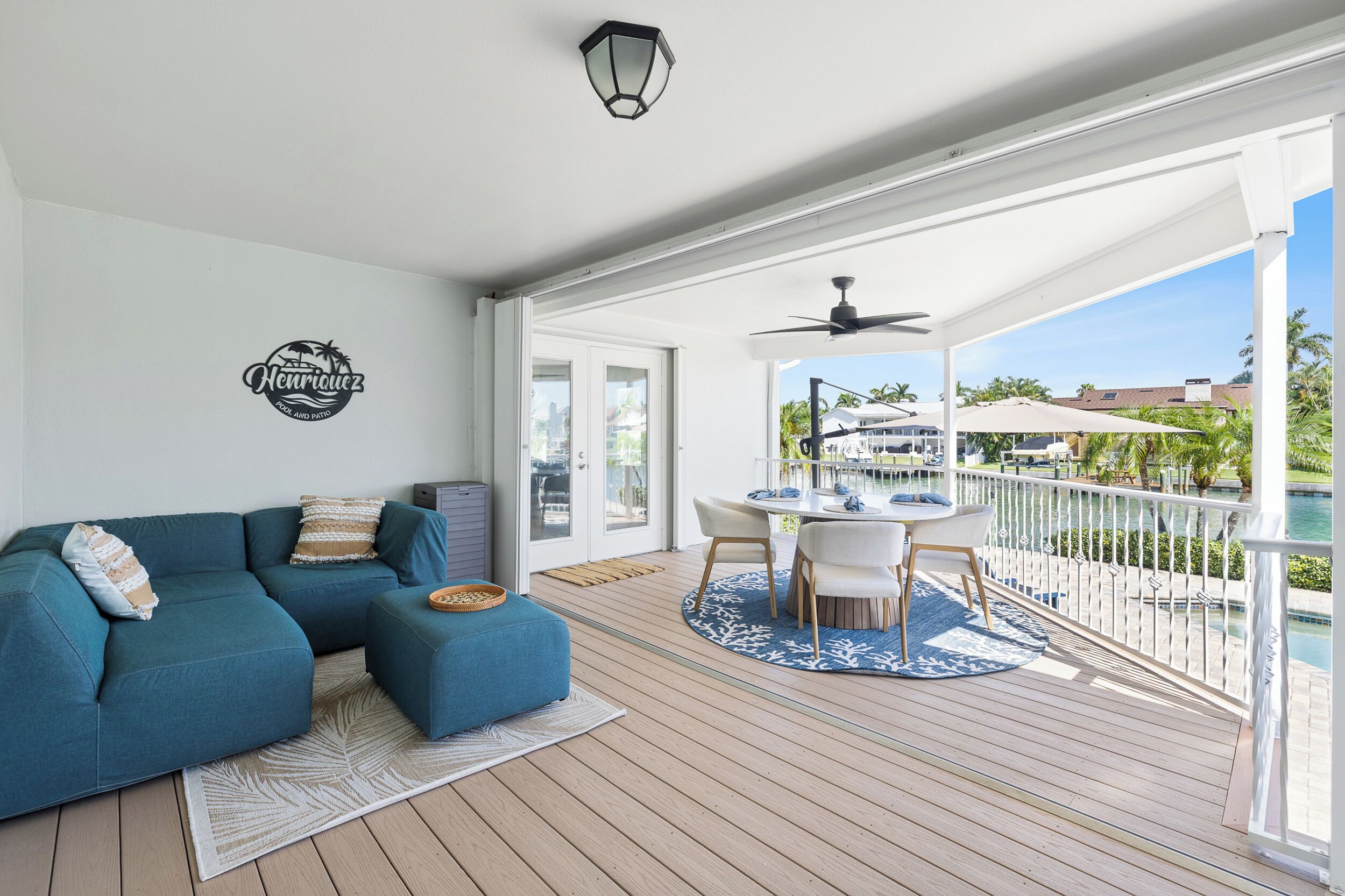 Ein überdachter Außenbereich mit einem blauen Sofa, einem Esstisch mit Stühlen, einem Deckenventilator und Blick auf ein Gewässer sowie Gebäude im Hintergrund.