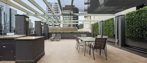 A rooftop terrace with a dining area, outdoor kitchen, and a view of surrounding buildings.