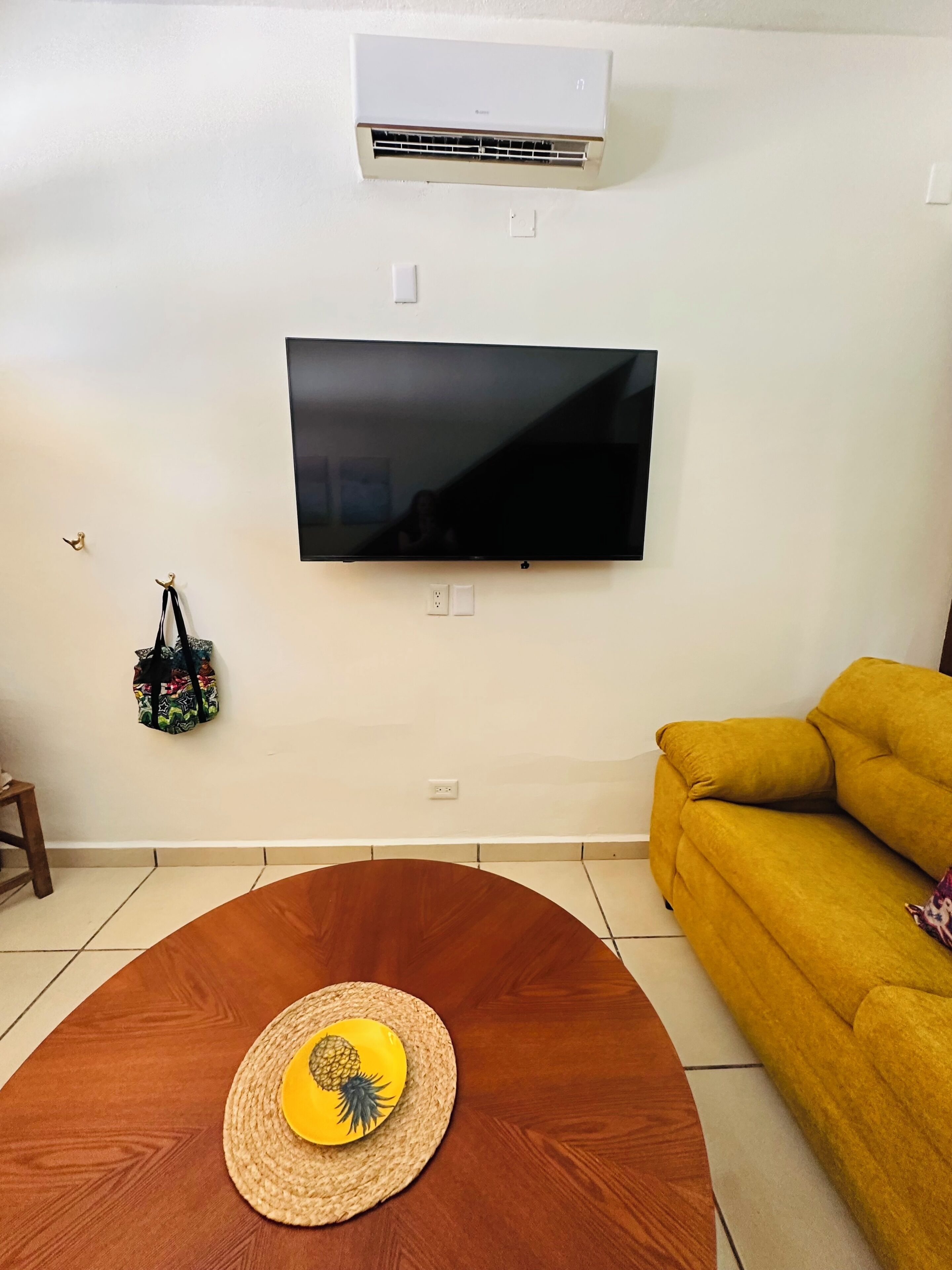 A living room with a flat-screen TV mounted on the wall, a yellow sofa, a round wooden coffee table with a pineapple design, and an air conditioning unit above.