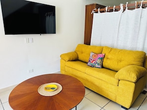 A room with a yellow sofa, a round wooden coffee table with a pineapple coaster, a flat-screen TV mounted on the wall, and a wooden cabinet with a white curtain.