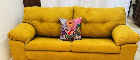 Un canapé jaune agrémenté d’un coussin coloré et une table ronde en bois ornée d’un motif représentant un ananas.