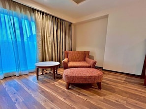 Una habitación con suelo de madera, un sillón marrón, una mesita redonda y una ventana con cortinas traslúcidas.