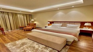 Quarto de hotel com uma cama grande, área de estar com uma poltrona e apoio para os pés, escrivaninha e janela com cortinas.