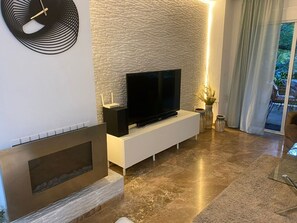 A modern living room with a flat-screen TV on a white cabinet, a stone-textured wall, a fireplace, and a view of the outdoors. - The Golf View Marbella, a peaceful golf retreat in Guadalmina (San Pedro Alcántara)