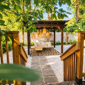 Eine Holz-Pergola mit einem weißen Sofa und einem gemusterten Teppich, umgeben von viel Grün und einem weißen Zaun.