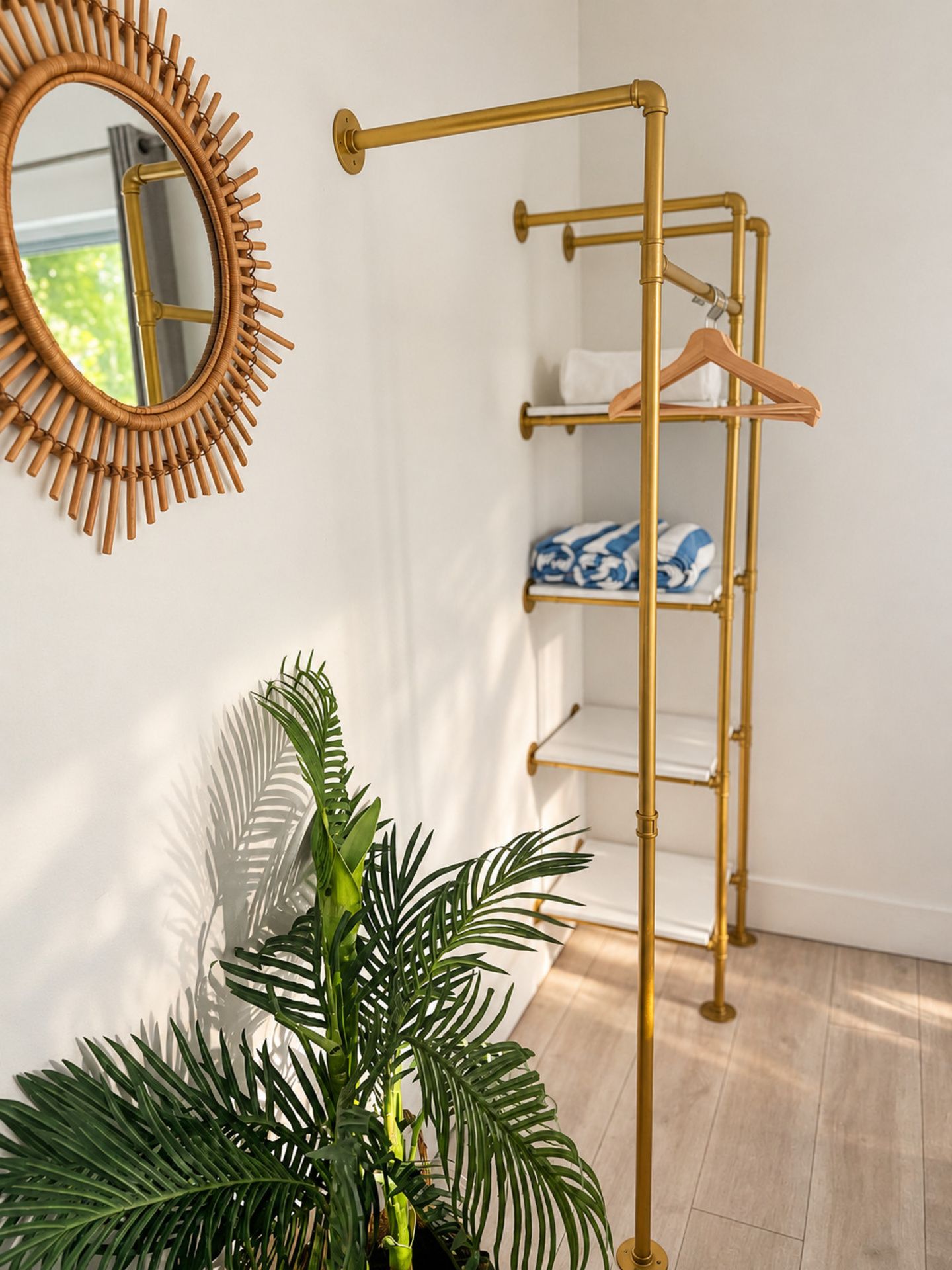 Une pièce avec une étagère métallique de couleur dorée adossée à un mur blanc, un miroir rond orné d’un motif en forme de soleil, et une plante en pot aux feuilles vertes.