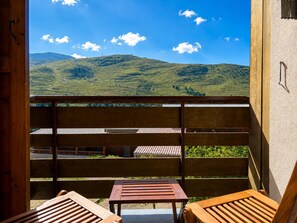 Ein Balkon mit Holzbalkonen und zwei Holzstühlen, der auf eine hügelige Landschaft mit blauem Himmel blickt.