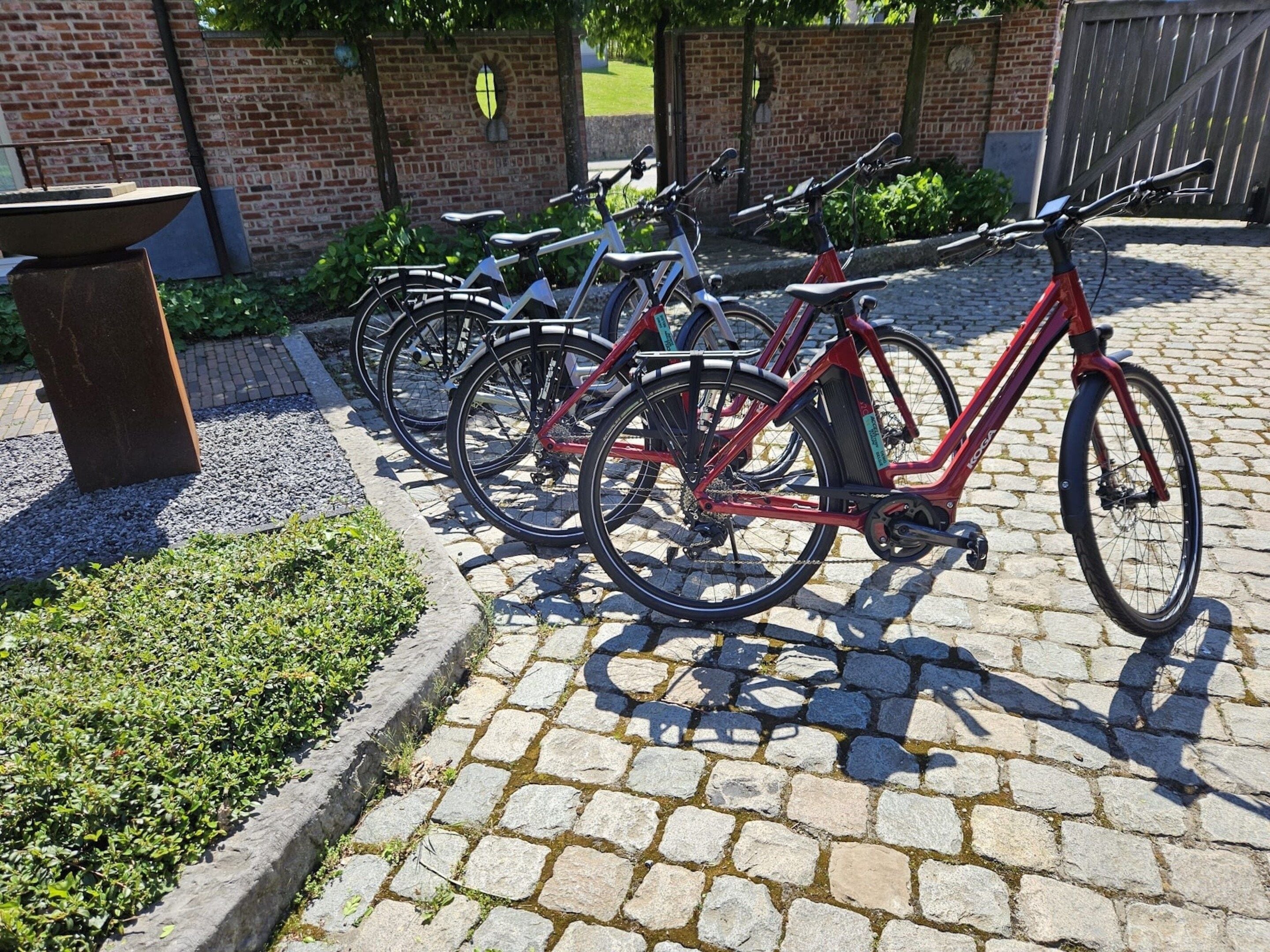 A row of bicycles parked on a cobblestone path with a brick wall and greenery in the background.