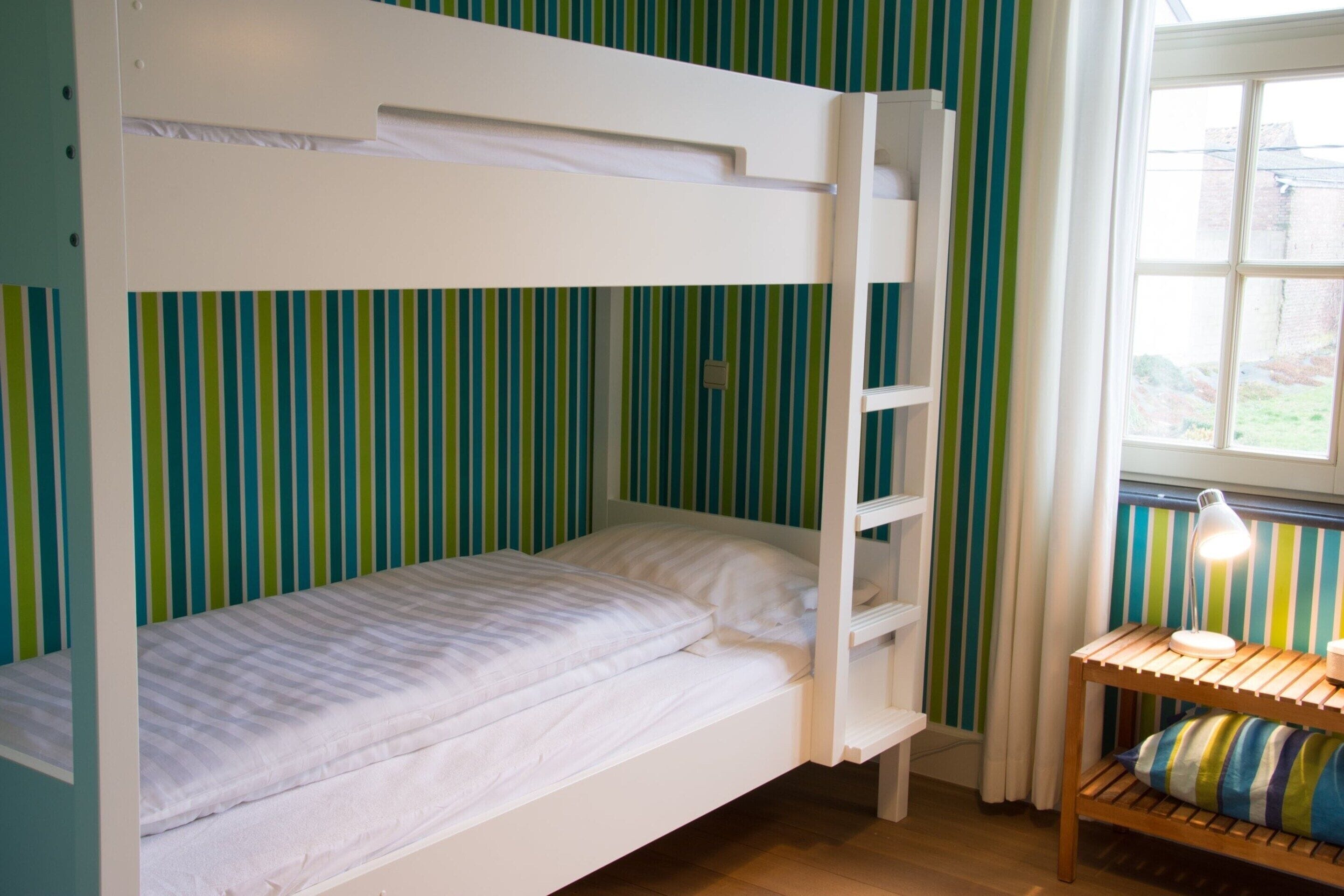 Una cama tipo litera con estructura blanca y estante de madera, una mesita de noche con lámpara y una ventana con cortinas.