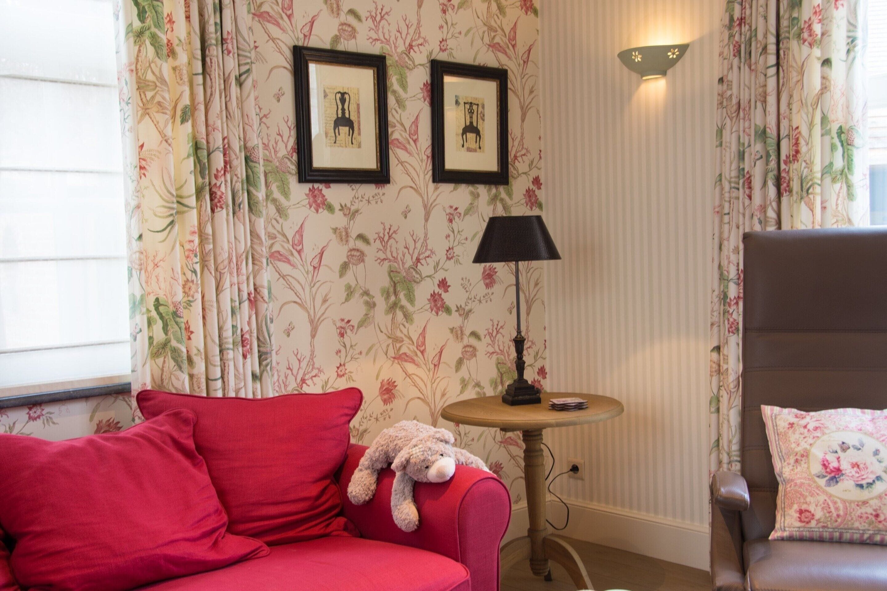 A room with a red sofa, a small round table with a lamp, a chair with a floral cushion, and two framed pictures on the wall.