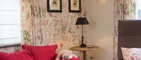 Una habitación con un sofá rojo, una mesita redonda con una lámpara, una silla con un cojín estampado y dos cuadros colgados en la pared.