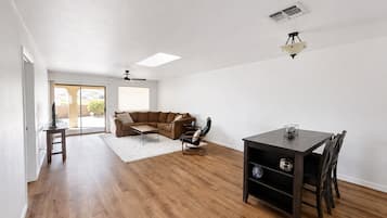 A spacious living area with a wooden floor, a brown sectional sofa, a dining table with chairs, and a ceiling fan.