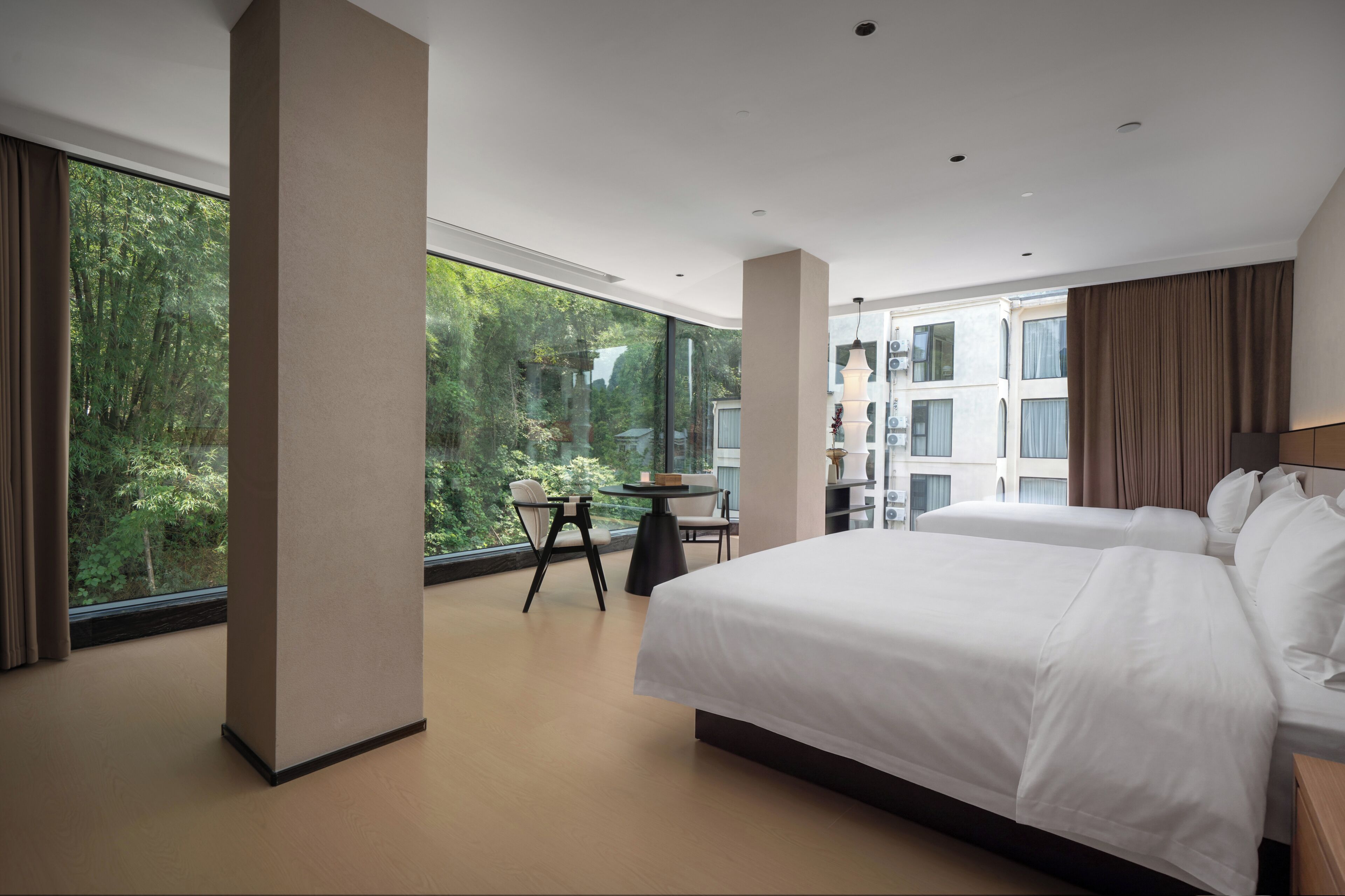 A hotel room with a large bed, a small table, and a chair. There is a view of greenery through the window.