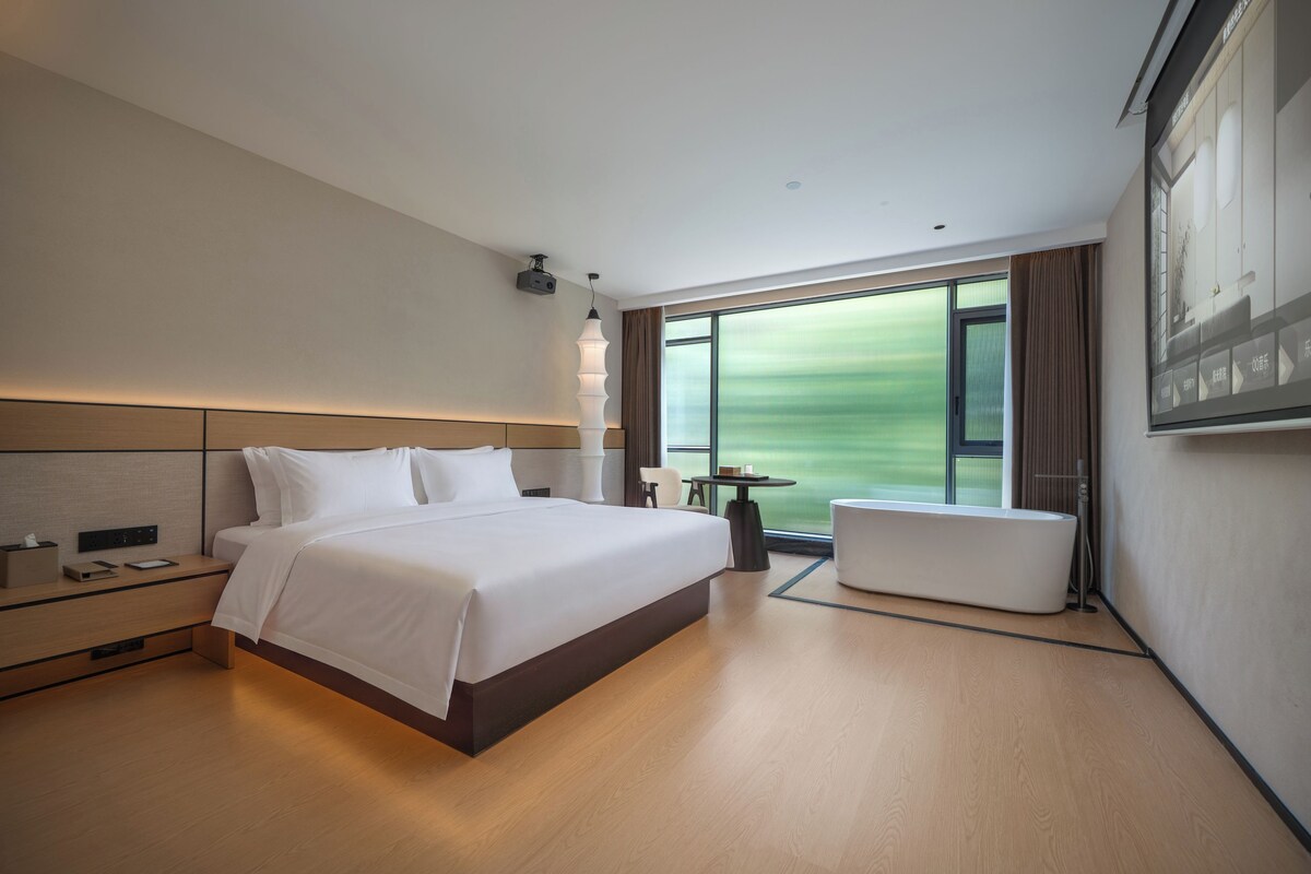 A modern hotel room with a large bed, a sitting area, and a view of a green landscape through the window.