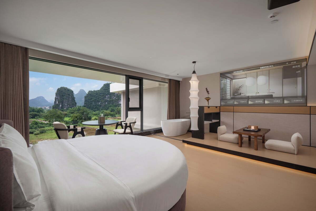 A modern hotel room with a large bed, a dining area, and a view of the mountains.