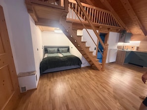Ein Schlafzimmer mit einer Holztreppe, die zu einem Schlafbereich im Zwischengeschoss führt, einem Bett mit dunkelgrüner Bettdecke und einer Küchenzeile mit Holzausstattung.