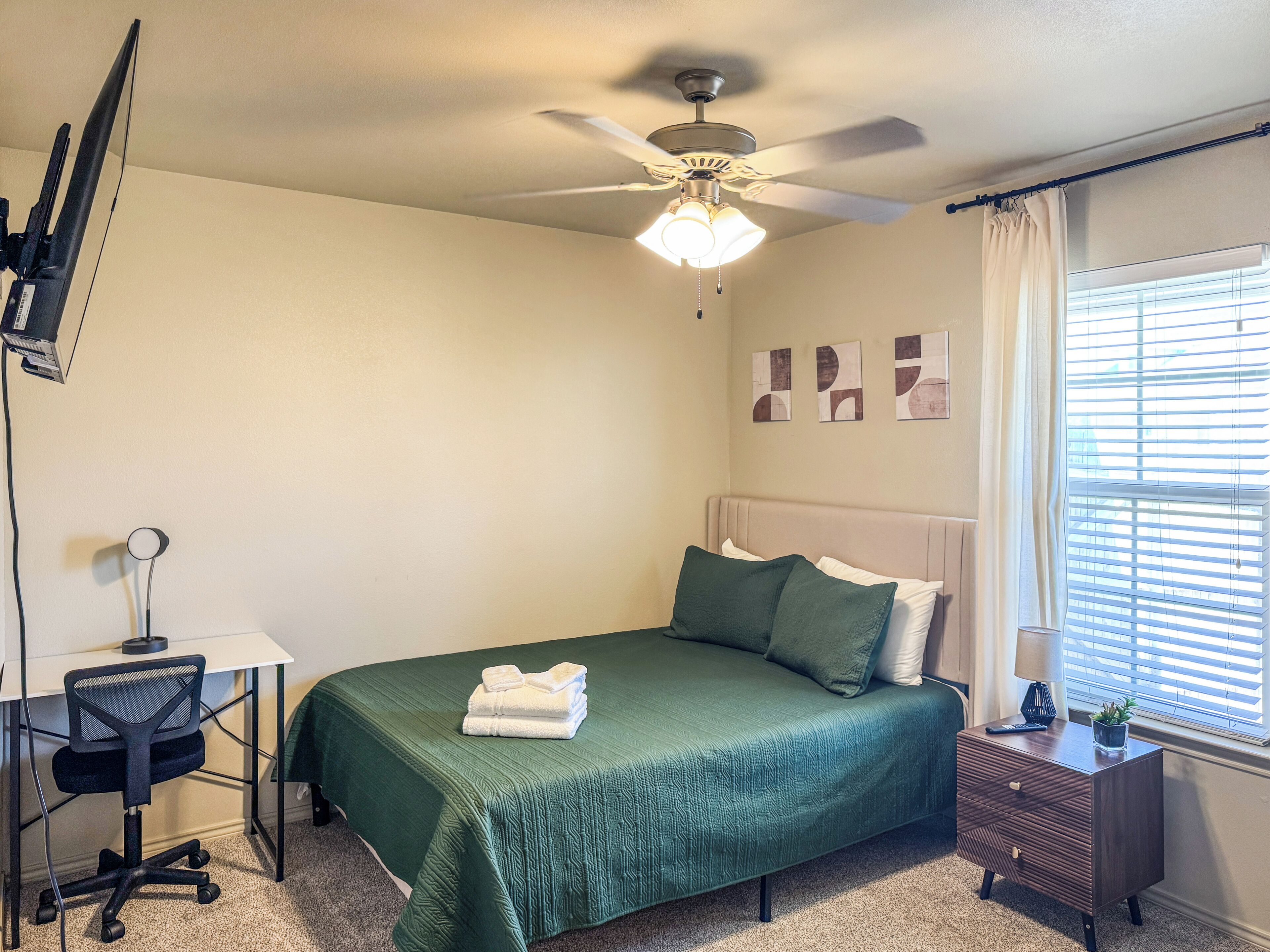 A bedroom with a bed, desk, chair, ceiling fan, and a window with blinds.