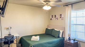 A bedroom with a bed, desk, chair, ceiling fan, and a window with blinds.