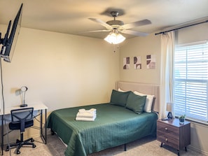 Un dormitorio con cama, escritorio, silla, ventilador de techo y ventana con persianas.
