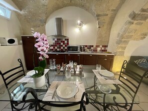A dining area with a glass table, chairs, and a potted orchid. There is a kitchen in the background with a microwave, stove, and tiled backsplash.