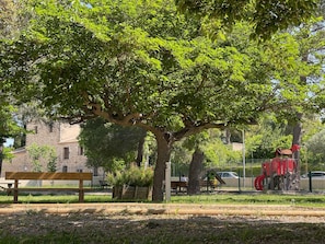 A park with a large tree, benches, a playground, and a stone building in the background.