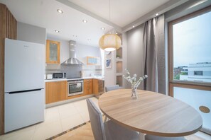A modern kitchen with a dining area, featuring a round wooden table and chairs, a built-in oven, and a large window with curtains. - Modern Suite Al. Wilanowska (Warszawa)