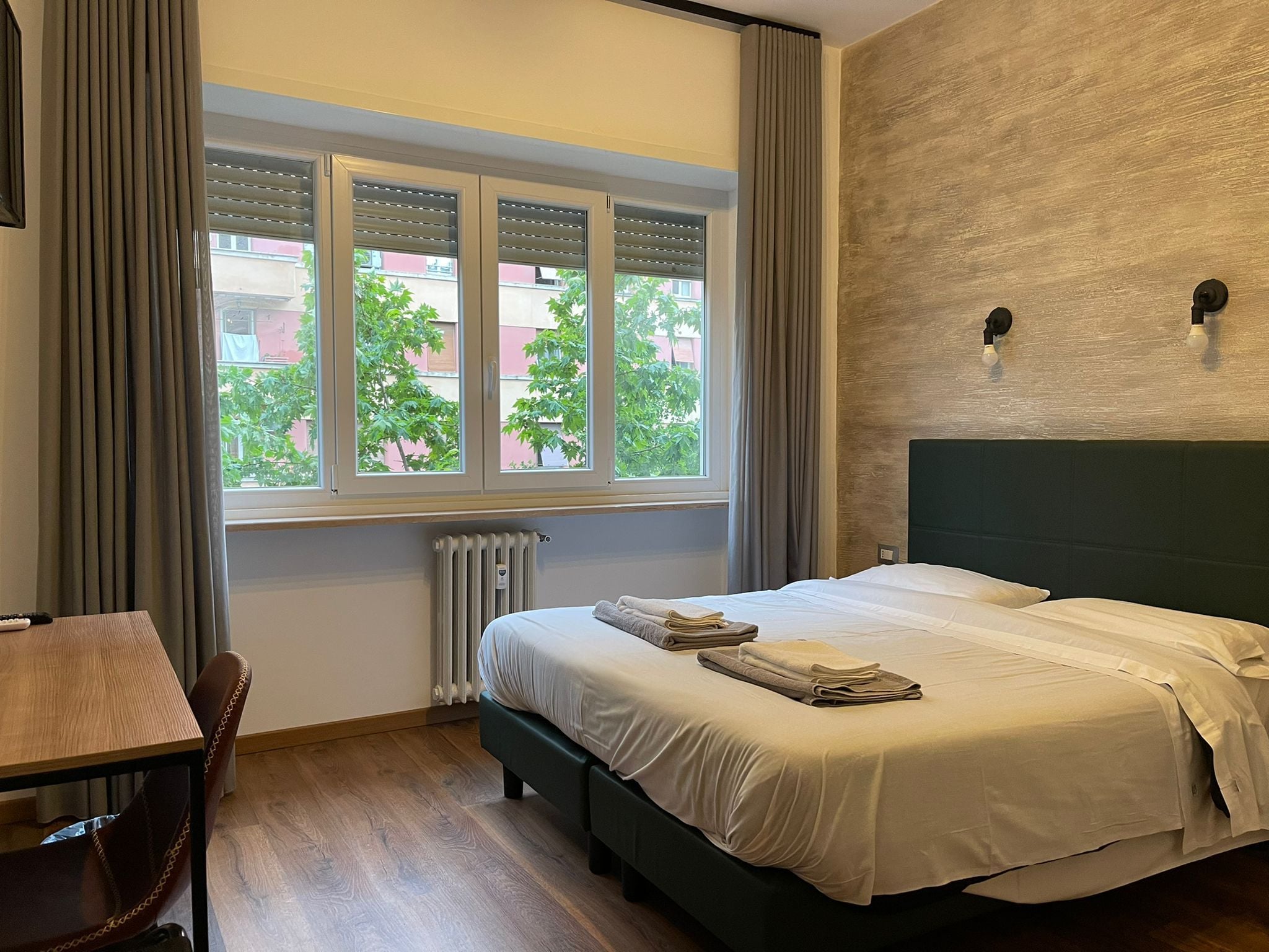 Habitació d'hotel amb un llit, una taula, una cadira, una finestra amb persianes i un radiador.