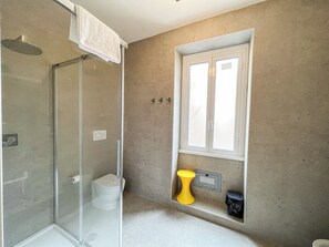 Un baño con cabina de ducha de vidrio, inodoro, ventana y un estante pequeño con un cubo de basura.