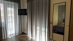 A room with a wooden floor, a bed, a TV mounted on the wall, and sheer curtains.