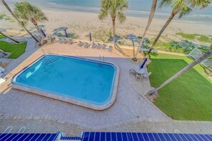 A rectangular swimming pool with a surrounding paved area, adjacent to a beach with palm trees and lounge chairs. - Welcoming 3-bedroom Beachfront Condo in Belleair Beach (Belleair Beach)