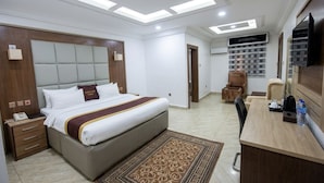 A hotel room with a bed, bedside table, desk, chair, and a wall-mounted TV. - Immaculate Platinum Luxury Resorts (Abuja)