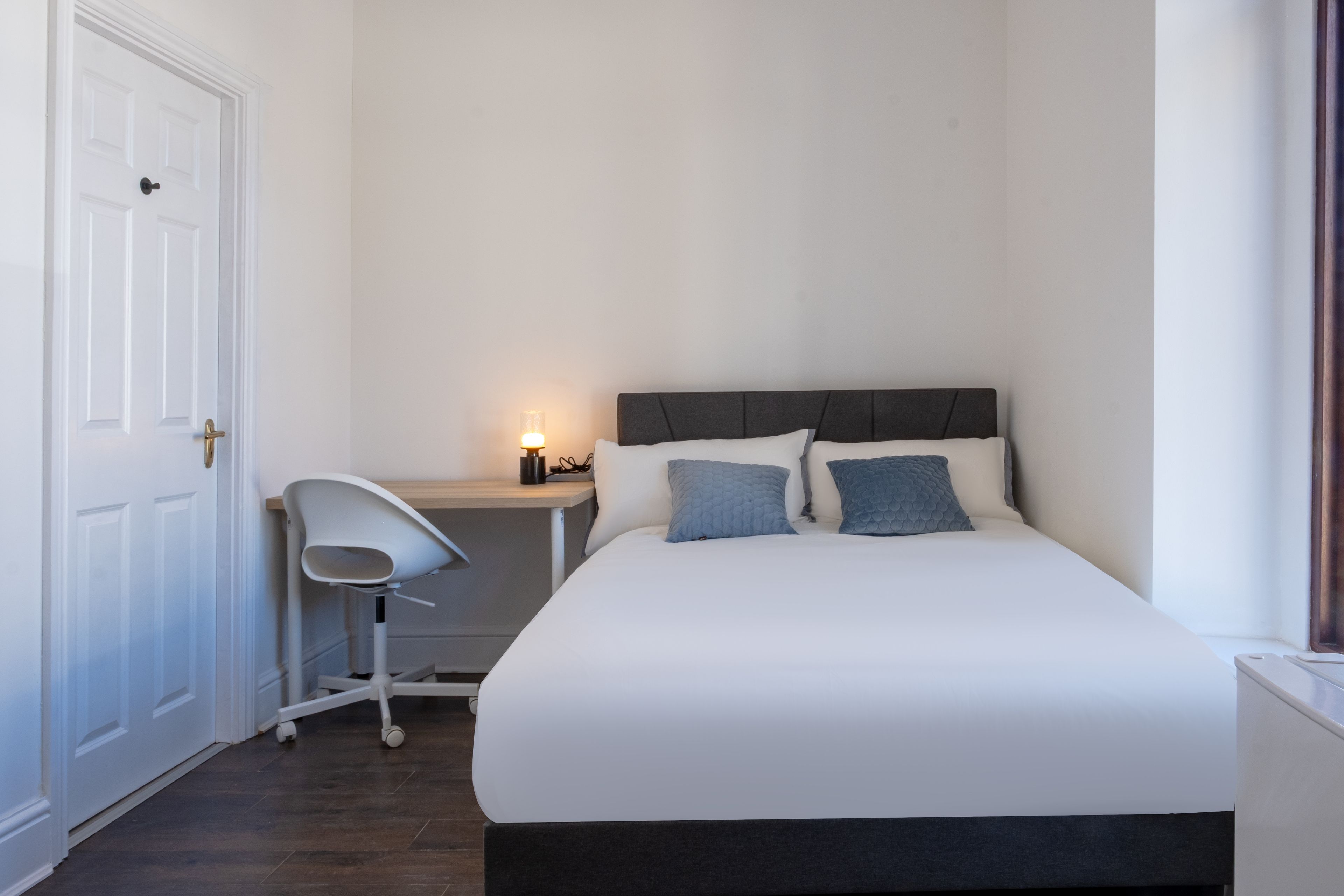 A bedroom with a bed, a chair, a bedside table with a lamp, and a door.