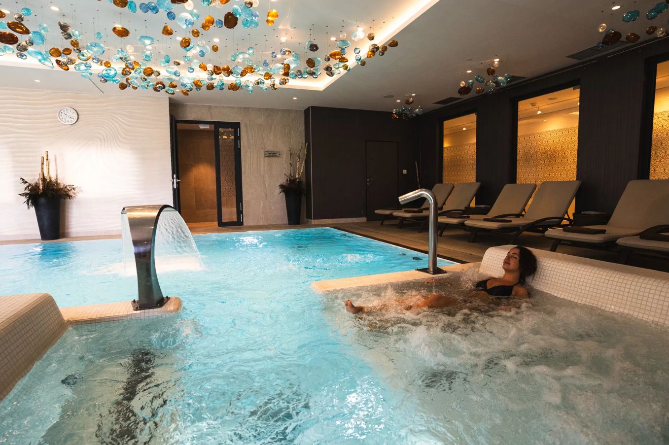 An indoor swimming pool with a person in a jacuzzi, lounge chairs, and a decorative ceiling.