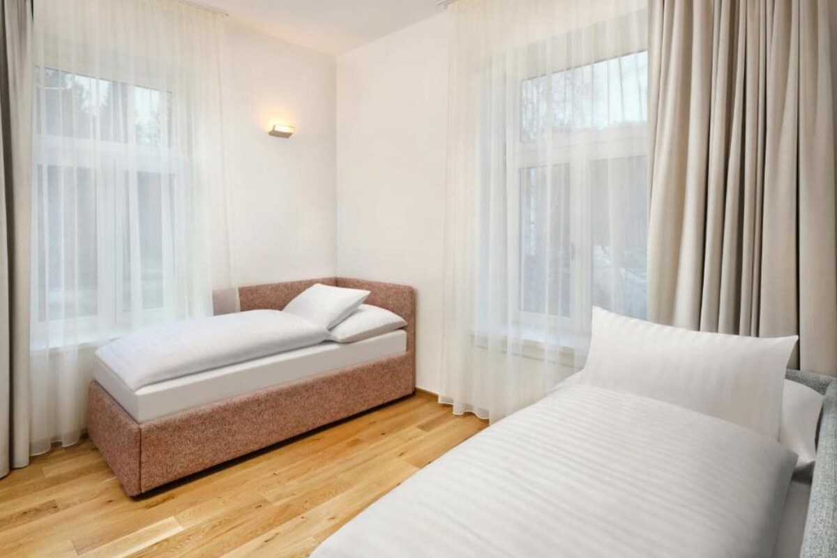 Ein Schlafzimmer mit einem Doppelbett, einem Einzelbett und einem Schlafsofa. Zwei Fenster mit leichtem Stoffvorhang, und eine Wandleuchte.